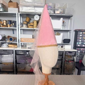 A pink velvet hat with a gold trim sits on a wooden stand in a cluttered craft room filled with shelves of supplies and materials.
