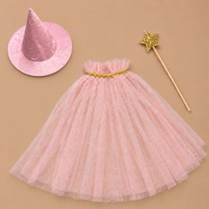 May include: A light pink cone-shaped hat with a wide brim, a matching light pink tulle cape featuring a gold-colored trim at the neckline, and a wooden wand topped with a gold glittery star. The set includes three distinct costume accessories.