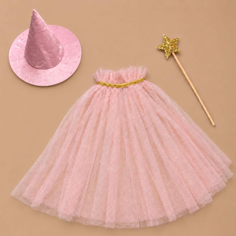 May include: A light pink cone-shaped hat with a wide brim, a matching light pink tulle cape featuring a gold-colored trim at the neckline, and a wooden wand topped with a gold glittery star. The set includes three distinct costume accessories.