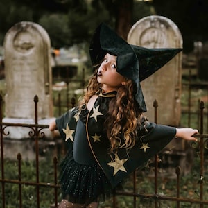 A young girl dressed as a witch, wearing a black outfit with gold stars, stands in front of a fence with two gravestones in a cemetery.