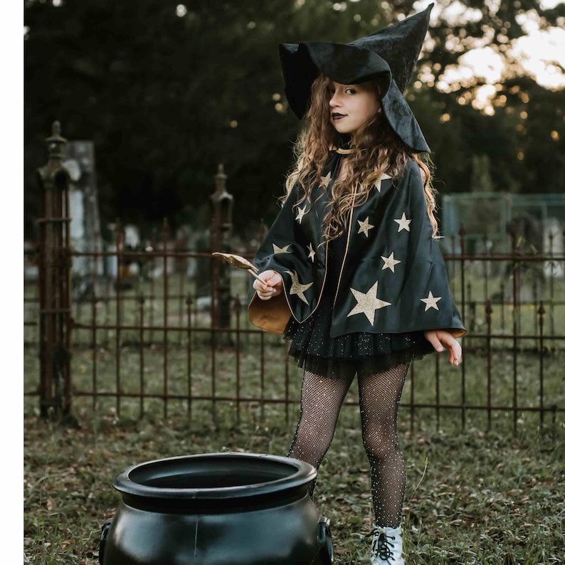 A young girl dressed as a witch, wearing a black outfit with gold stars, stands next to a large black cauldron in a garden setting.