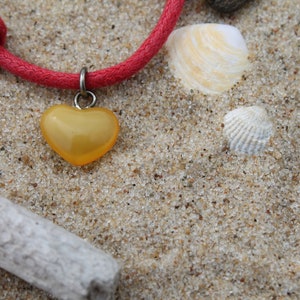 May include: A red cord bracelet with a yellow heart charm on a sandy background. The heart charm is made of glass and has a shiny finish.