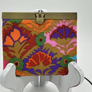 May include: A square fabric coin purse featuring a vibrant, multi-colored floral pattern in shades of orange, purple, pink, green, and brown. The purse is secured with a rectangular, antique brass-toned metal frame and a central push-lock clasp.