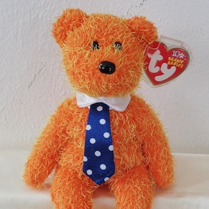 May include: Orange Beanie Baby with a blue and white polka dot tie and a white collar. The bear is sitting on a white surface. The tag says "10th Beanie Babies".
