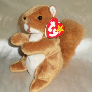 May include: A brown and white plush squirrel with a long, bushy tail. The squirrel is sitting up with its paws in front of it. It has a red heart-shaped tag attached to its ear that says "Beanie Babies".