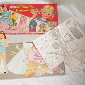 May include: A vintage paper doll set with a stand-up doll and multiple outfits. The box features a red background with the text "MAGIC Stay On Dresses" and a picture of a girl holding a sign that says "LOOK".