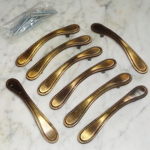 Puede incluir: Conjunto de ocho tiradores de puerta de armario de oro antiguo con borde de cuentas y diseño curvo.