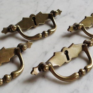 May include: Four antique brass cabinet door pulls with a keyhole design. Each pull has a decorative, ornate design and a curved handle.