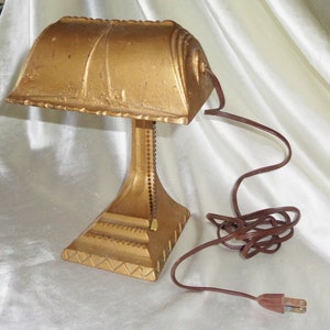 May include: A gold-colored metal desk lamp with a curved shade and a chain to adjust the height. The lamp has a brown cord and plug.