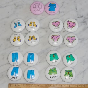 以下が含まれることがあります： さまざまな種類の服のカラフルなイラストが描かれた、白い丸いボタンが16個セットになっています。靴下、下着、ジーンズ、ボクサーパンツ、パジャマ、シャツなどがあります。ボタンには服の名前が書かれています。