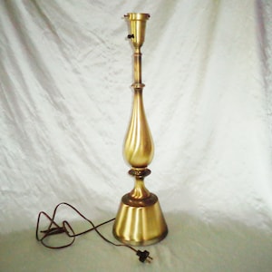 May include: A tall, gold-toned table lamp with a slender, curved base and a simple, round shade holder. The lamp has a brown cord and a black on/off switch.