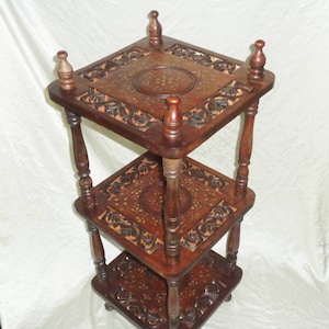 May include: A three-tiered dark brown wooden stand featuring square shelves with carved floral borders and central circular brass inlay patterns. The structure is supported by four turned wooden pillars and topped with four small decorative finials.