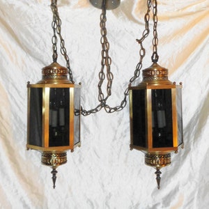 May include: A pair of brass and glass pendant lights with black glass shades hanging from chains. The lights have a decorative design around the top and bottom of the glass shades.