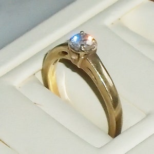 May include: A gold-colored ring with a single, clear gemstone. The ring is set in a classic style, with a simple band and a raised setting for the stone. The ring is displayed in a white jewelry box.