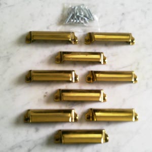 May include: A collection of eight polished brass drawer pulls, arranged in rows on a white marble surface. A small bag of silver screws is in the upper right corner. Each pull is rectangular with rounded edges.