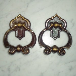May include: Two antique bronze drawer pulls with intricate designs. Each pull has a decorative top, a central oval handle, and scrolled details. The pulls are likely metal and designed for furniture, with a vintage aesthetic.