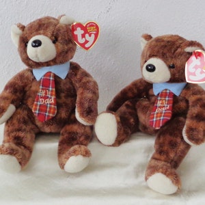 Puede incluir: Dos osos de peluche marrones que llevan camisas azules y corbatas de cuadros rojos y negros. Las corbatas tienen el texto "#1 Dad" bordado. Los osos están sentados sobre una superficie blanca.