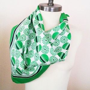 May include: A green and white patterned scarf draped on a mannequin. The scarf features a repeating floral design with green and black accents. The scarf has a green border and is made of a silky material.