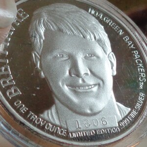 May include: A silver coin featuring a portrait of a smiling man. The coin is inscribed with "BRETT FAVRE", "ONE TROY OUNCE", "NO.4 GREEN BAY PACKERS", "999 FINE SILVER", and "1806 LIMITED EDITION".