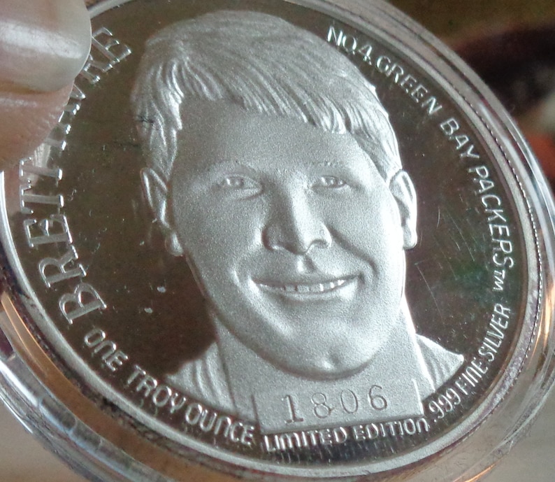 May include: A silver coin featuring a portrait of a smiling man. The coin is inscribed with "BRETT FAVRE", "ONE TROY OUNCE", "NO.4 GREEN BAY PACKERS", "999 FINE SILVER", and "1806 LIMITED EDITION".