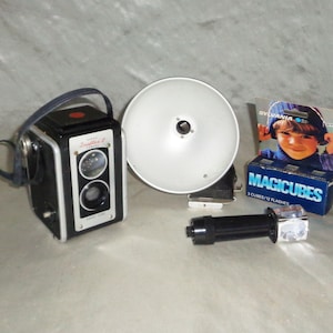 May include: A black and silver vintage camera with a flash attachment, a flash cube, and a box of flash cubes labeled "Sylvania Magicubes 3 Cubes/12 Flashes".