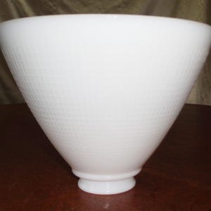 May include: A white, cone-shaped glass lampshade with a textured diamond pattern. The lampshade has a wide opening and a small, rounded base. The shade is made of opaque glass and is sitting on a dark wooden surface.