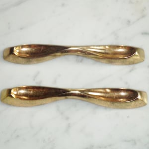 Könnte beinhalten: Zwei goldfarbene Metall-Schrankgriffe mit einem gedrehten, geschwungenen Design.