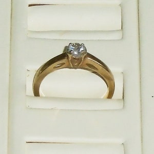 May include: A gold engagement ring with a single, clear gemstone. The ring is set in a classic, four-prong setting. The band has a simple, elegant design. The ring is displayed in a white jewelry box.