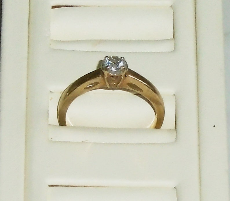 May include: A gold engagement ring with a single, clear gemstone. The ring is set in a classic, four-prong setting. The band has a simple, elegant design. The ring is displayed in a white jewelry box.