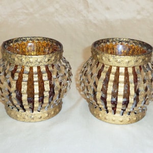 May include: Two gold metal candle holders with a clear amber glass insert. The candle holders have a decorative, geometric design.