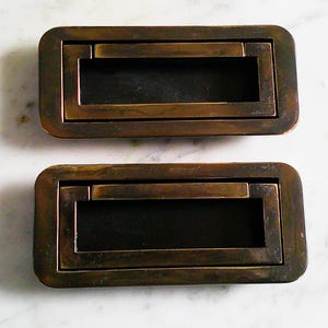 Puede incluir: Dos tiradores rectangulares de cajón de latón antiguo. Cada tirador tiene un asa empotrada y un interior oscuro. Los tiradores se colocan sobre un fondo de mármol blanco, mostrando su estética vintage y envejecida.