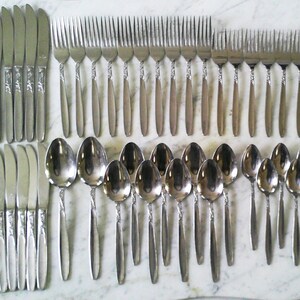 May include: A collection of stainless steel silverware, including knives, forks, and spoons. The knives and forks have decorative handles, while the spoons have a twisted design. The silverware is arranged on a light-colored surface.