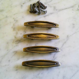 Puede incluir: Cuatro tiradores de cajón de latón antiguo con un diseño curvo, junto con un pequeño montón de tornillos. Los tiradores tienen un acabado cálido y envejecido, lo que sugiere un estilo vintage. Ideal para la restauración de muebles.