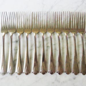 May include: A collection of ten silver forks arranged in a row. Each fork has a long, slender handle with a decorative design near the base and four tines. The forks are placed on a white and gray marble surface.