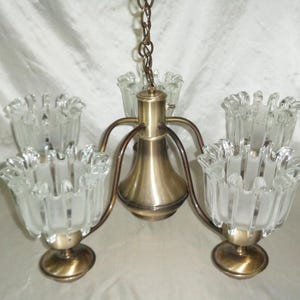 Może przedstawiać: Zabytkowy mosiężny żyrandol z pięcioma kloszami z matowego szkła. Żyrandol ma centralną mosiężną oprawę z zakrzywionymi ramionami podtrzymującymi szklane klosze. Klosze mają falisty brzeg i są zawieszone na łańcuchu.