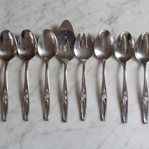 May include: A set of eight silver-toned serving utensils, including spoons, a serving fork, and a slotted spoon. The utensils have a floral design on the handles.