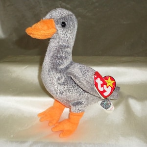 May include: Grey and orange plush goose with a Ty Beanie Baby tag attached. The tag is red with a white star and the words "Beanie Babies" in white.