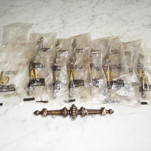 May include: Six packs of gold-toned cabinet hardware with the brand name "bewith" printed on each pack. A single piece of hardware is shown outside of the packaging. The hardware is a decorative, ornate, gold-toned cabinet pull.