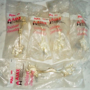 Puede incluir: Cinco paquetes individuales de tiradores de armario dorados con un diseño floral. Cada paquete tiene el texto "AJAX" y "Cabinet Jewelry" impreso en él.