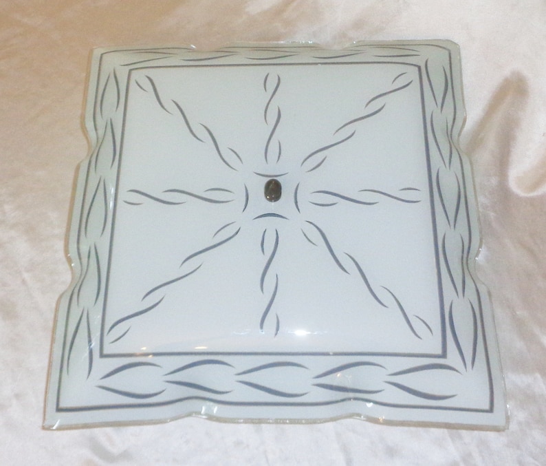 May include: A white frosted glass ceiling light fixture with a square shape and a decorative border of wavy lines. The center of the fixture has a circular design with a small metal knob.