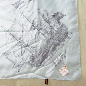 May include: A light blue silk scarf featuring a detailed black and white illustration of a sailing ship. The scarf has a tag that reads "From the House of Berkum Riff". The scarf is square and has a sewn edge.