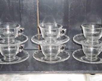 May include: Six clear glass teacups and saucers with a delicate scalloped edge. The cups and saucers are stacked in pairs.