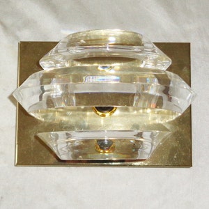 May include: A layered crystal wall sconce with a brass backing plate. The sconce has a geometric shape and is designed to cast a warm, ambient light.