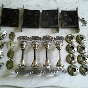 May include: A collection of vintage door hardware, featuring four black metal mortise locks, glass doorknobs with silver bases, oval escutcheons, and brass rosettes. Small screws are scattered around the components.