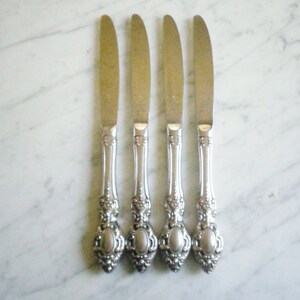 Puede incluir: Cuatro cuchillos de cena de plata con mangos ornamentados. Los mangos tienen un diseño decorativo con un óvalo en relieve en el centro.