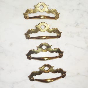 Puede incluir: Cuatro tiradores de armario dorados con diseños ornamentados. Cada tirador tiene una forma curva y un centro decorativo.