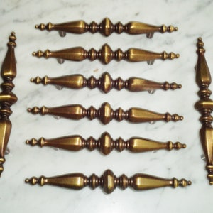 May include: A collection of antique brass drawer pulls and decorative vertical handles. The drawer pulls have a central bulbous design with tapered ends. The handles have a similar design, with a series of stacked, decorative elements. The hardware is arranged on a white marble surface.