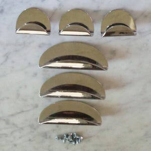 May include: A set of six polished chrome half-moon drawer pulls with a curved design. The pulls are arranged in two rows, with a small pile of screws at the bottom. The hardware is reflective and sits on a light-colored surface.