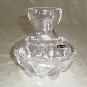 May include: A clear glass vase with a textured, faceted design. The vase has a wide base and a narrow neck. The label on the vase reads "Form Lars Hellsten".