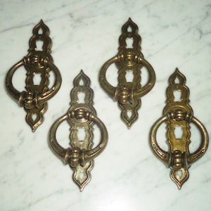 Könnte beinhalten: Vier antike Schubladengriffe aus Messing mit Ringgriffen. Jeder Griff hat eine verzierte Rückplatte mit Schlüssellochdesign und dekorativen Details. Die Griffe sind auf einer weiß-grauen Marmoroberfläche angeordnet.
